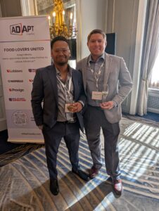 Two men in suits pose with drinks at a conference, standing next to an ‘Adapt Media’ banner that reads ‘FOOD LOVERS UNITED.’ The ornate chandelier and windowed room are in the background.