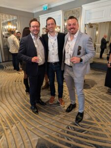 Three men in suits posing together at a formal networking event in an elegant banquet room, all wearing lanyards with name badges.