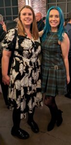Two women posing together at an indoor event smiling at the camera one in a black and white floral dress the other in a green plaid dress with blue hair